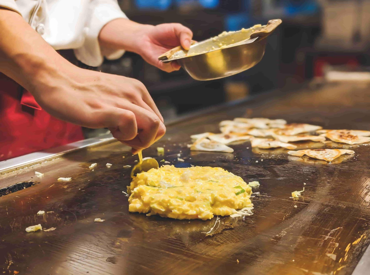 A close-up image of Okonomiyaki being cooked on a grill.