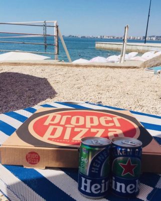 A proper pizza box and two Heineken cans placed on a blue and white striped sunchair on a beach.